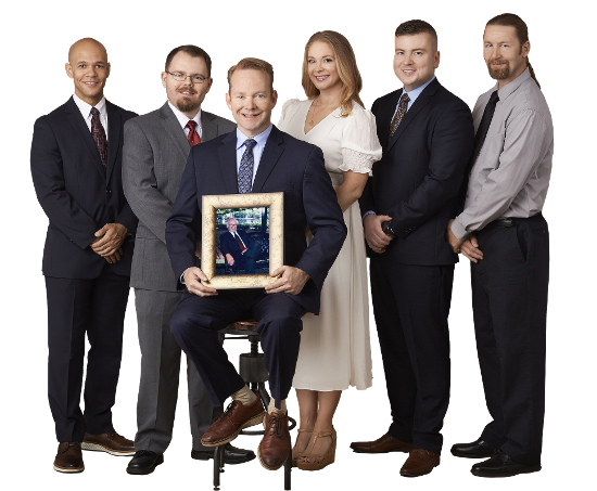 Six people in business attire pose in a studio, one seated holding a framed award or photo.