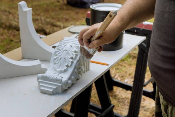 A person paints a white ornate decorative molding with a paintbrush on a white surface, outdoors.
