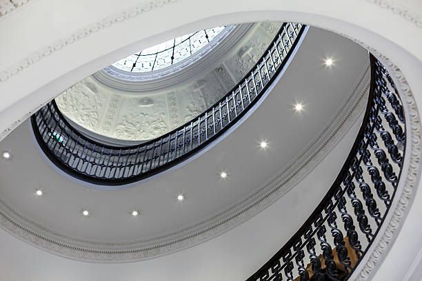 View from below of a white spiral staircase with black railings, lit by spotlights and a skylight.
