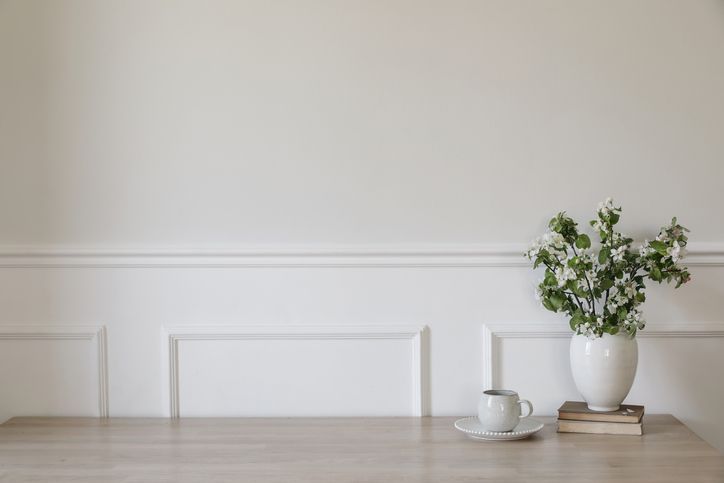 A white vase with greenery sits on a stack of books beside a cup and saucer on a light-colored wooden table against a white wall.