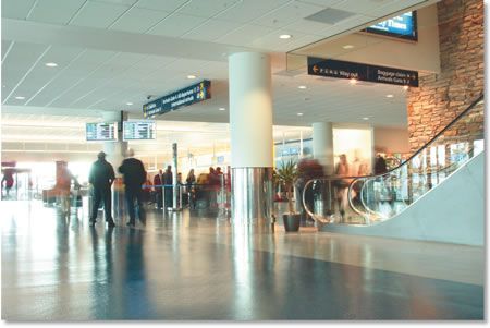 Interior of an airport terminal with passengers, signage, and an escalator. Shiny floor reflects the light.