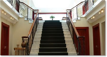 A grand staircase with black carpet, marble steps, and red doors. A vase and table sit on the left.