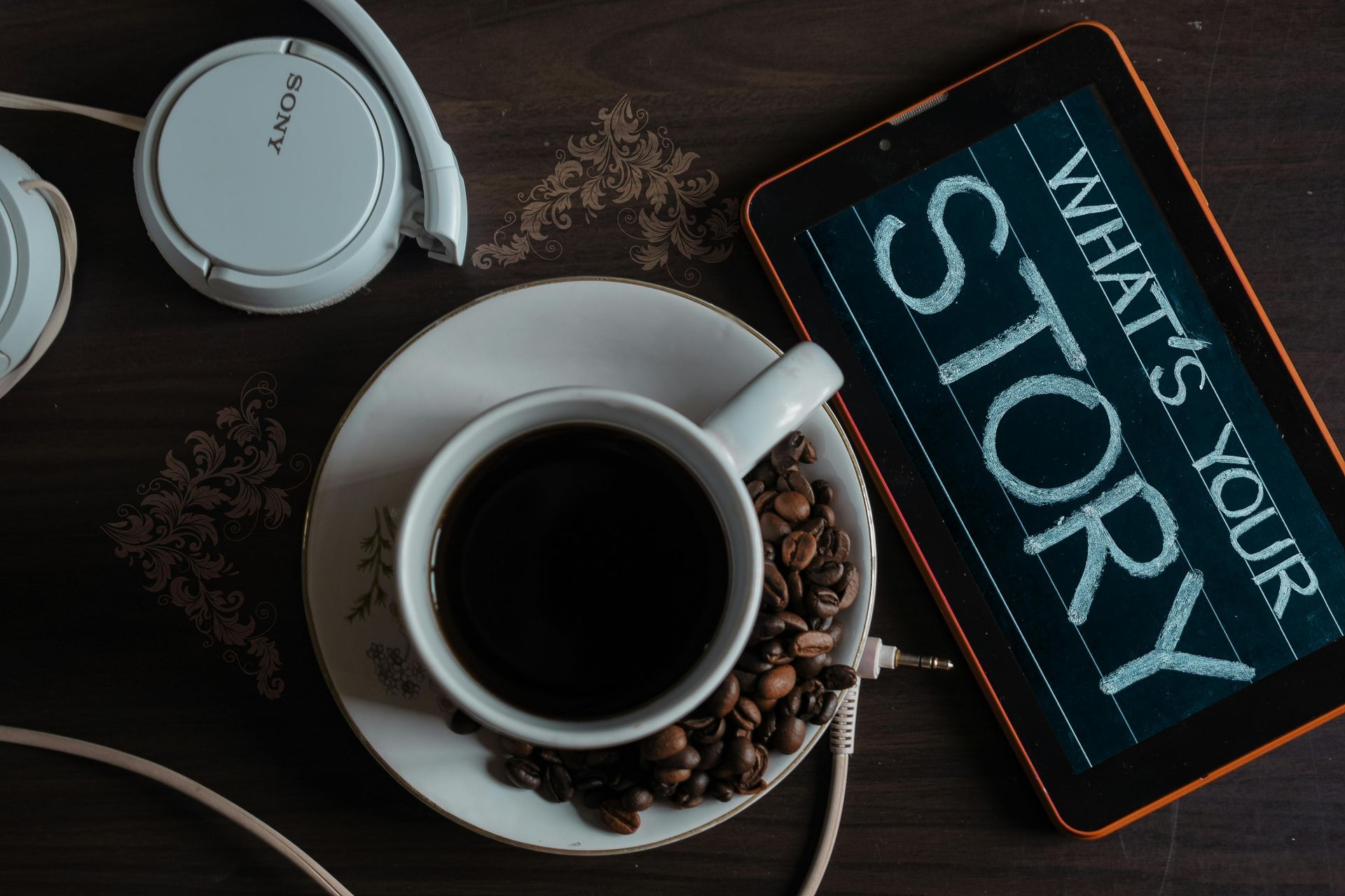 A cup of coffee on a saucer next to a tablet and headphones.