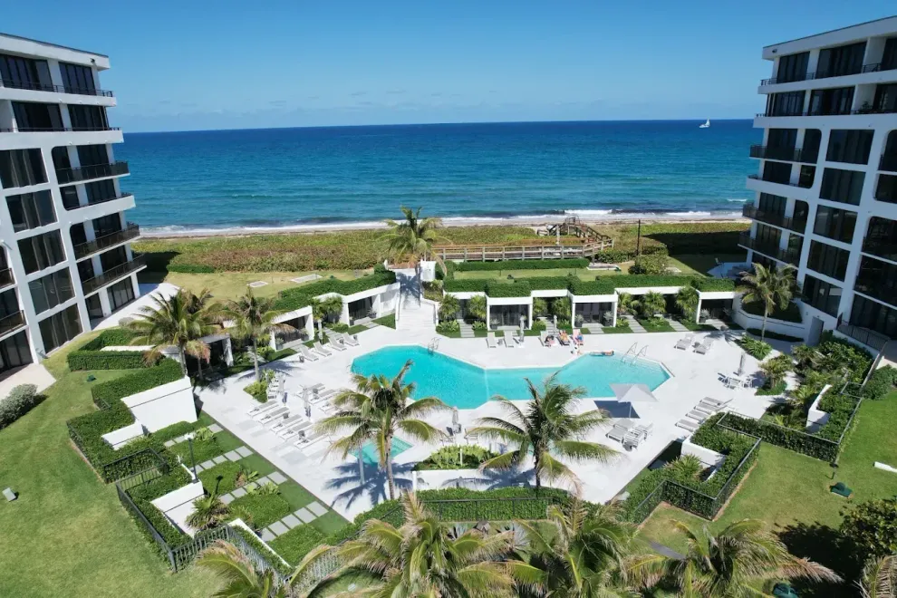 Oceanfront condo complex with pool, palm trees, and blue sky.