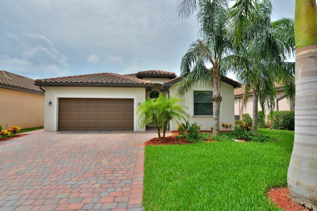 Beige stucco house with brown garage door, brick driveway, green lawn, palm trees.
