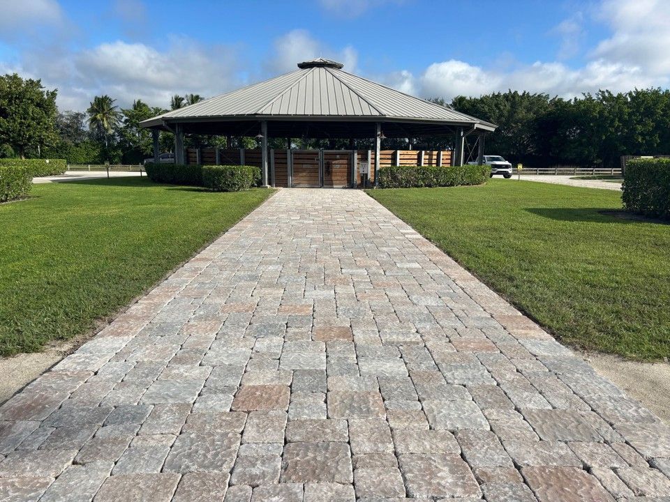 Brick pathway leading to a pavilion with a metal roof, surrounded by green grass and shrubs.