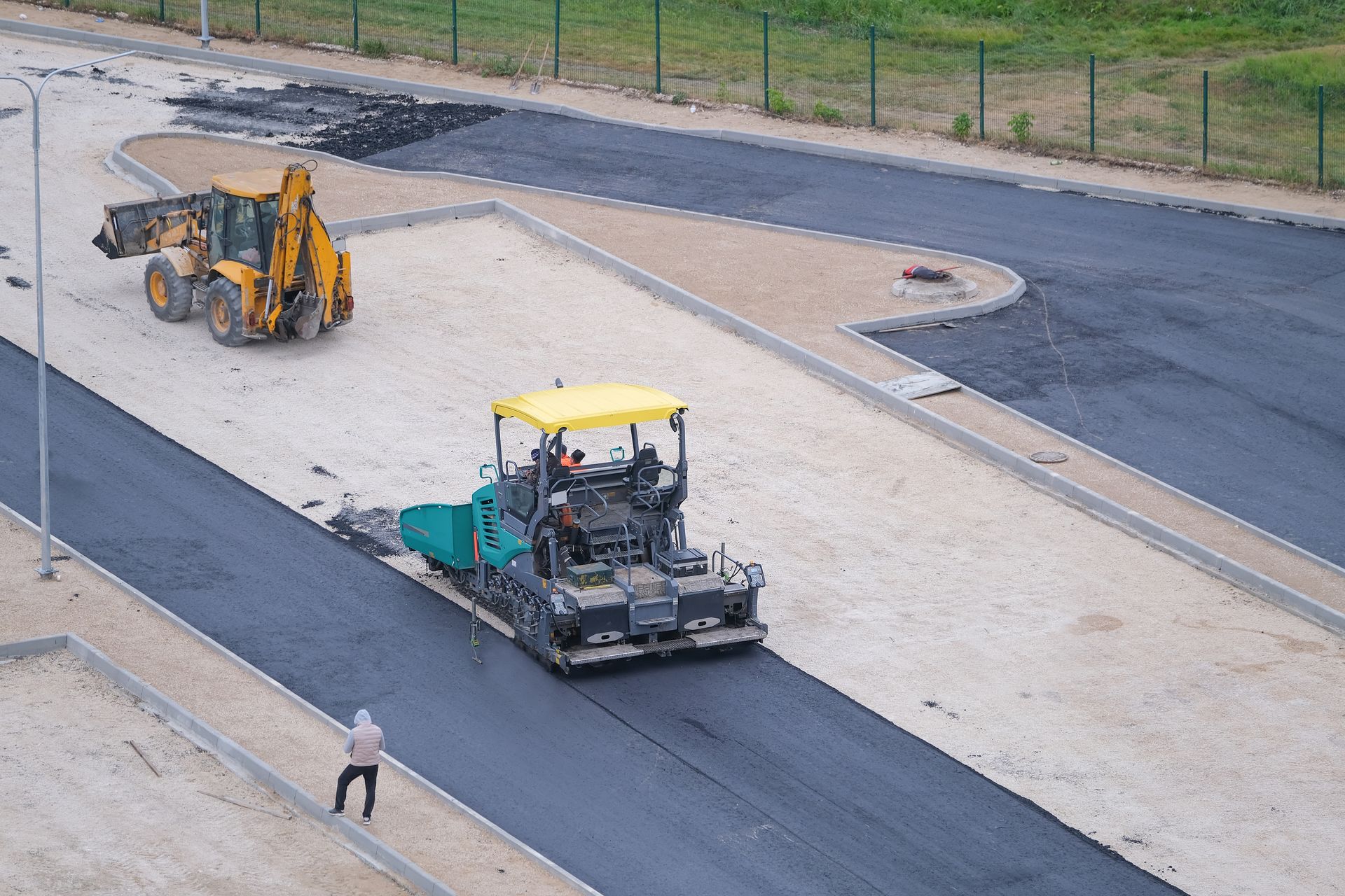 Roadwork crew paving a road with a yellow machine and a small excavator on a closed street