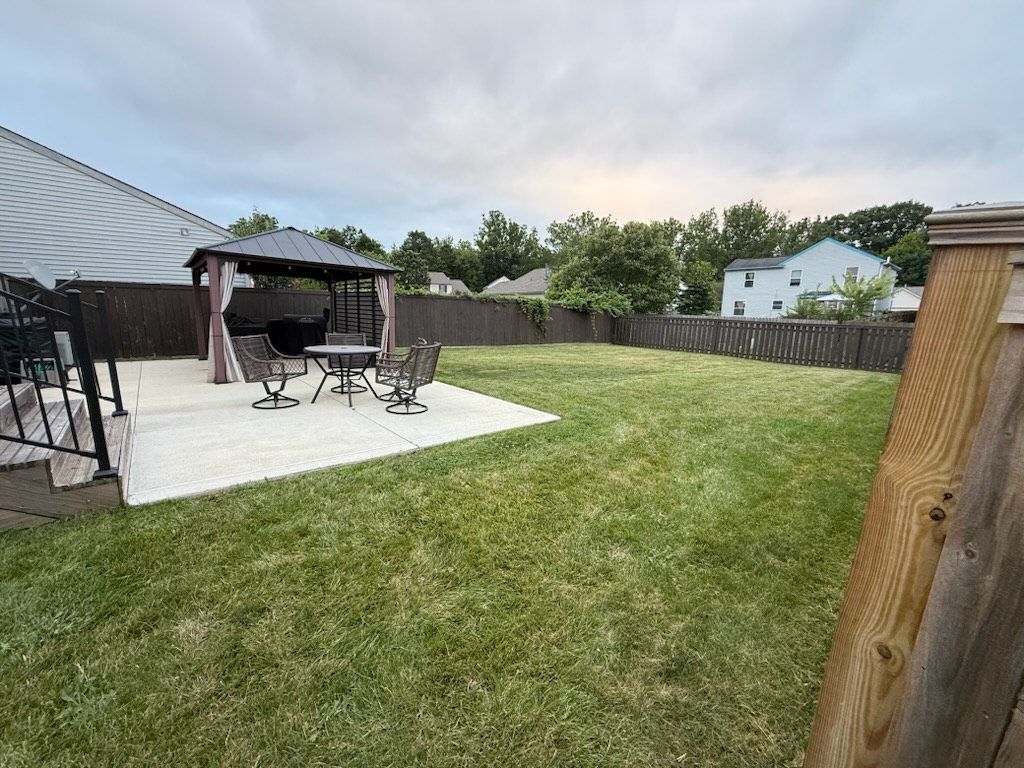 Backyard with patio, gazebo, table, chairs, and a wooden fence. Green grass and overcast sky.
