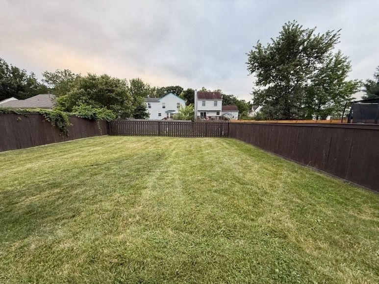 Lawn of green grass in backyard with brown wooden fence, houses, and trees in the distance under a cloudy sky.