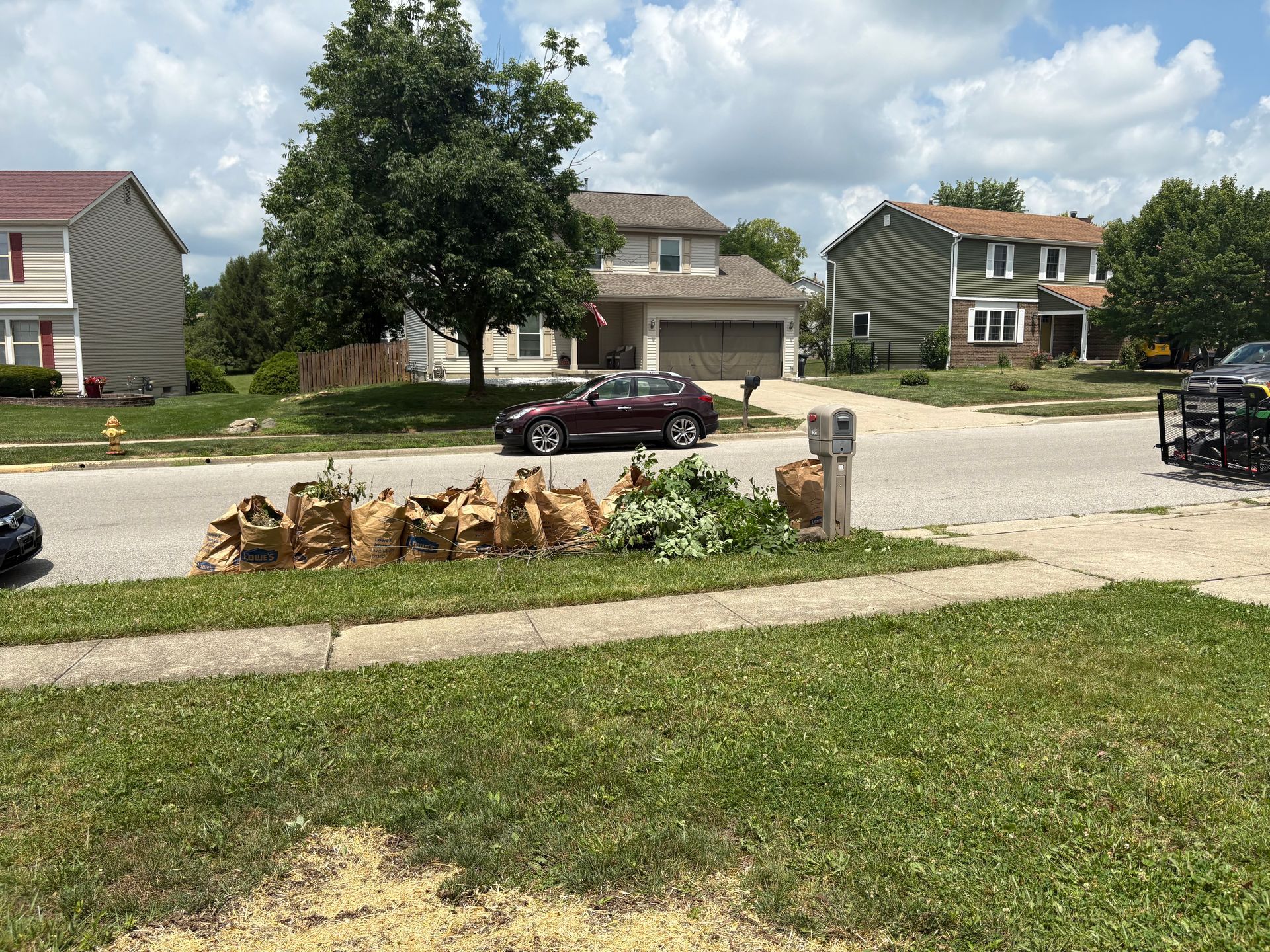 A residential street with houses, a car, tree stumps, and a lawn.