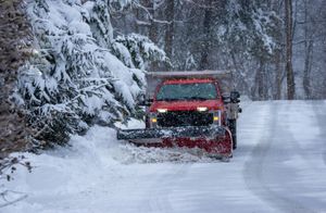 Red snowplow clearing snow from a road lined with snow-covered trees during a snowfall.