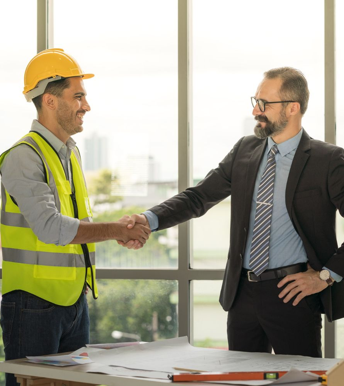 Construction worker and businessman shaking hands in an office, with plans on the table.