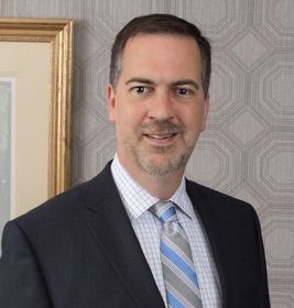 Man in a dark suit and blue striped tie, standing indoors against patterned wallpaper.