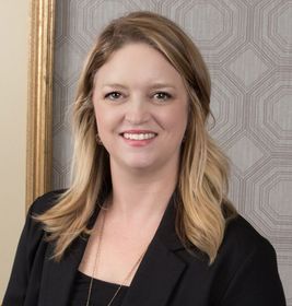 Smiling woman in a black blazer against a patterned wall.