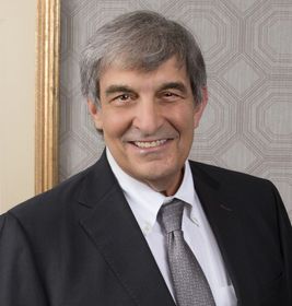 Smiling man in a dark suit and gray tie against patterned wallpaper.