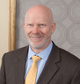 Smiling bald man in a suit and yellow tie against a patterned wall.