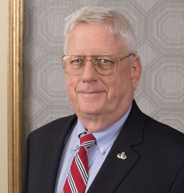 Portrait of a man in a navy suit, red striped tie, and glasses against a patterned wall