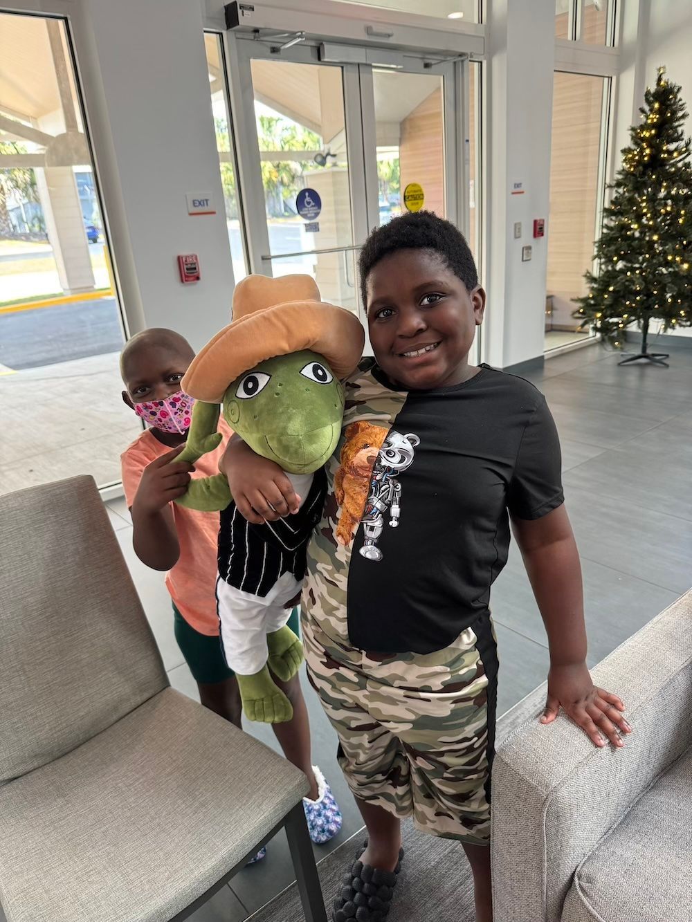 Two children with a stuffed animal, one wearing a cowboy hat. Indoors with a Christmas tree.