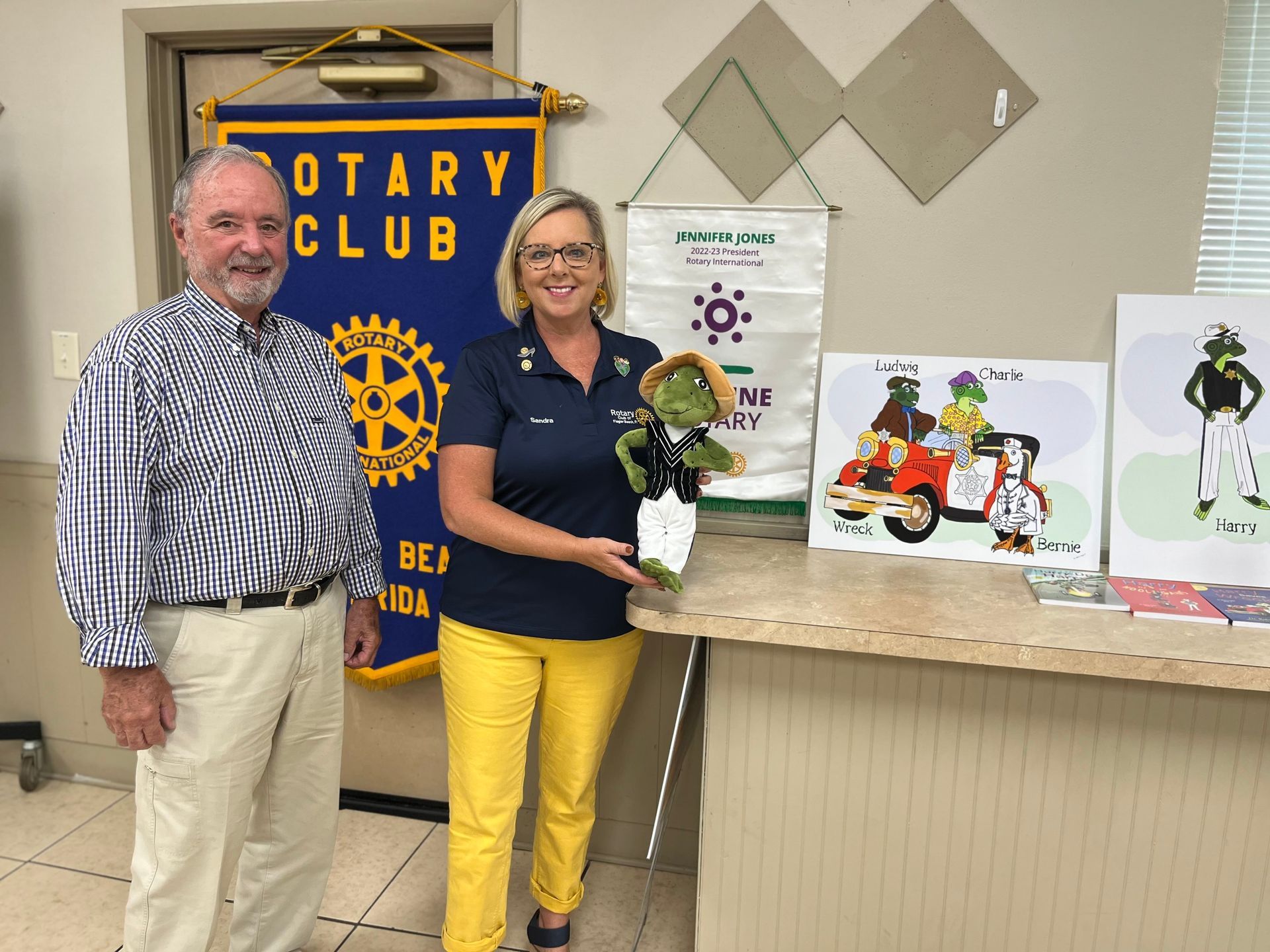 Man and woman holding a toy reptile and standing near Rotary Club banner and drawings.