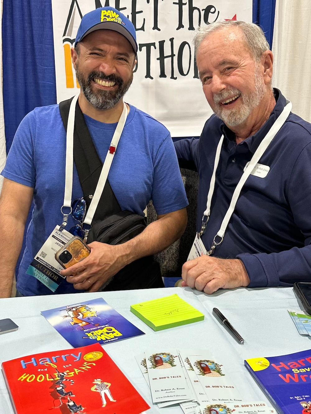 Two men smiling at a booth. One wears a blue cap and shirt, the other a blue polo. Books are displayed.