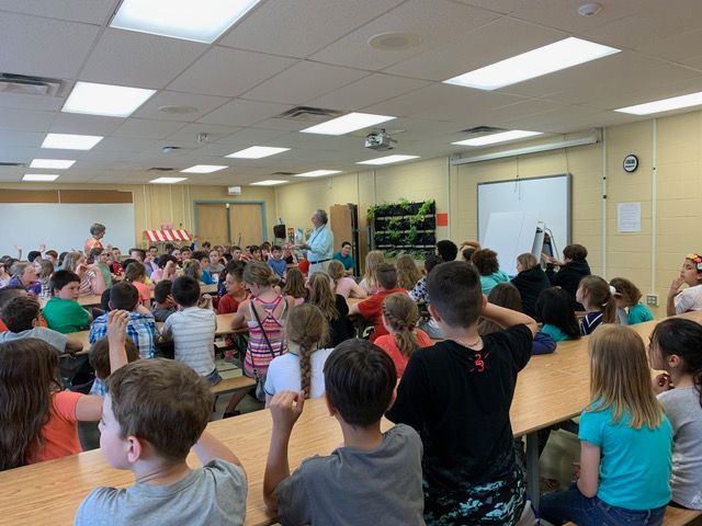 Students in a cafeteria listen to a presentation. Tables and a whiteboard are visible.