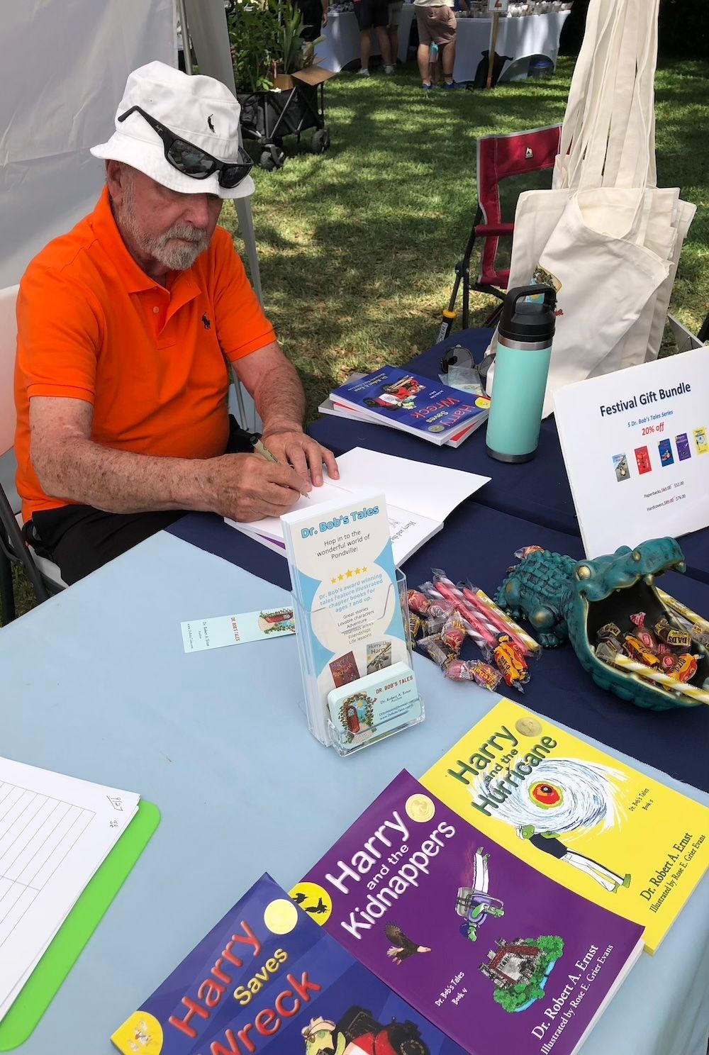Man at a table with books. He wears a hat and sunglasses, writing. Books, brochures, and a water bottle are on the table.