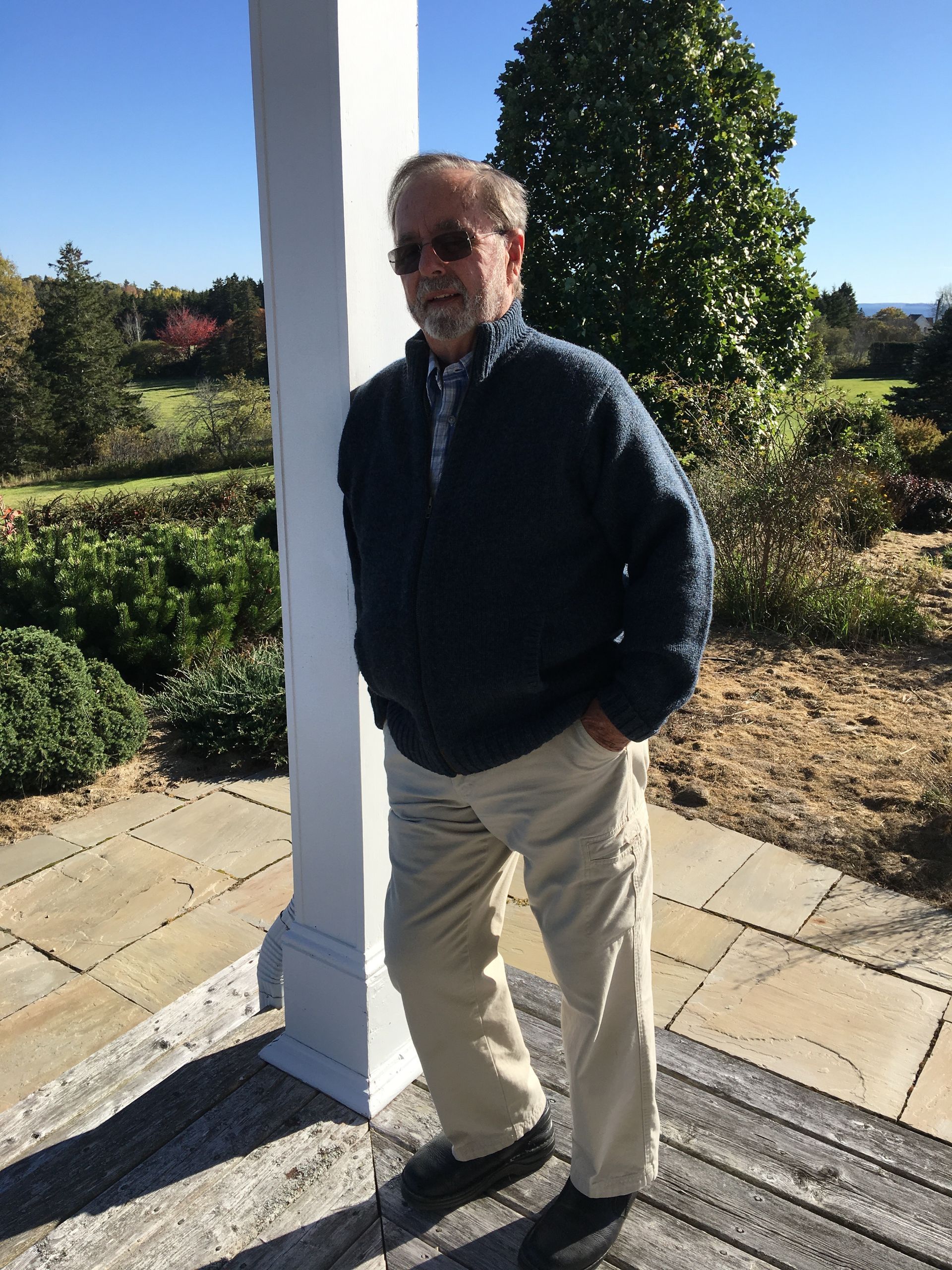 Man in blue sweater and khaki pants stands on a stone patio, leaning on a white column.