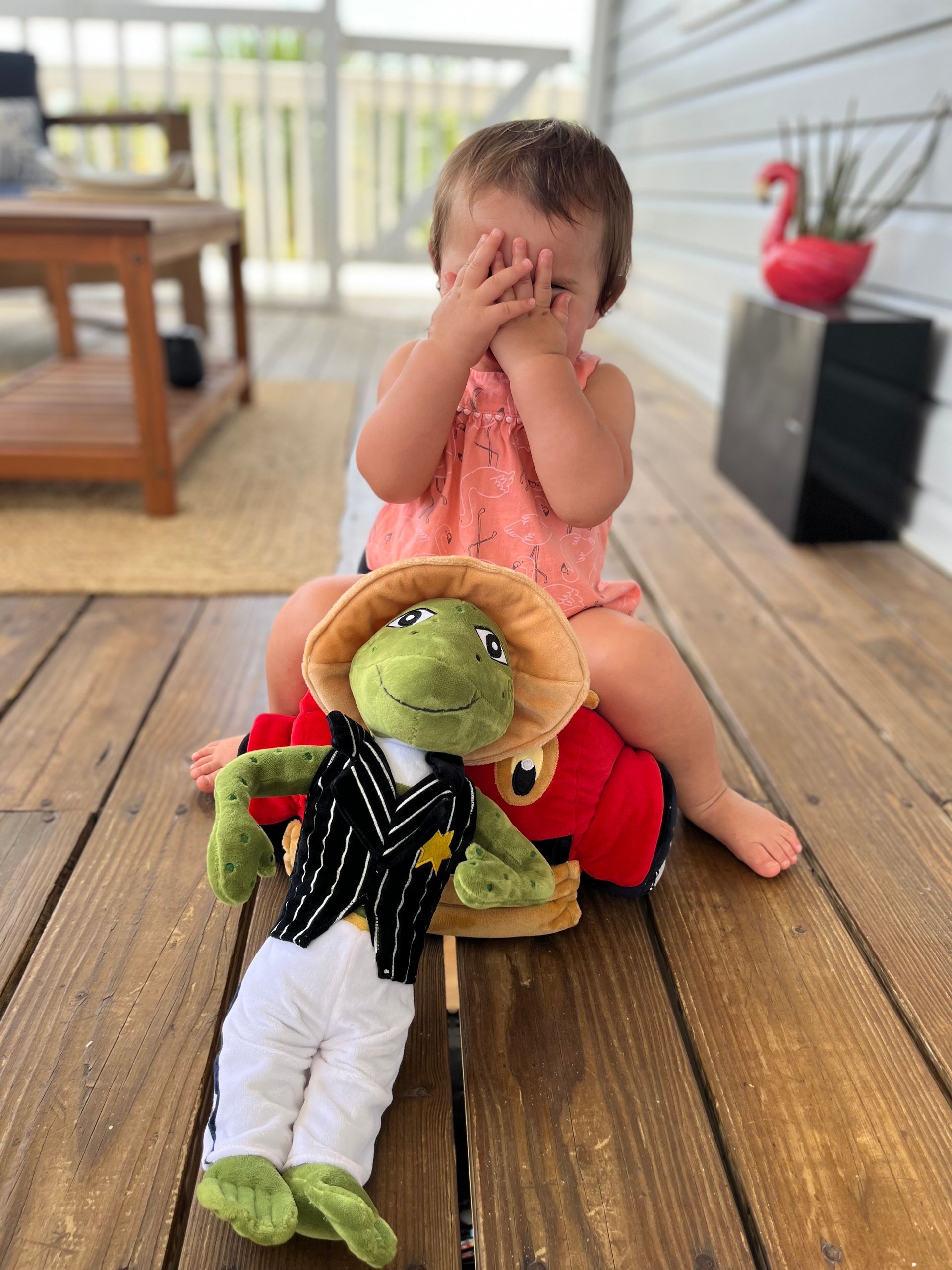 Child covering eyes, sitting on a red toy, with a frog toy in front of them on a wooden porch.