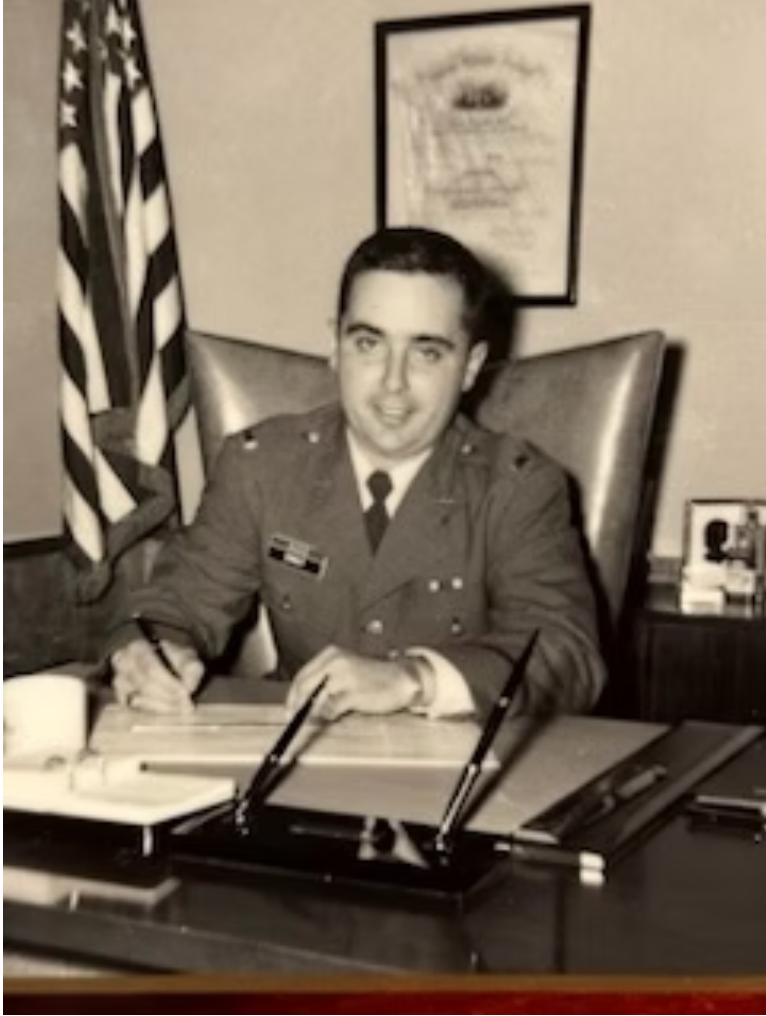 Man in military uniform sits at a desk, writing. An American flag is behind him.