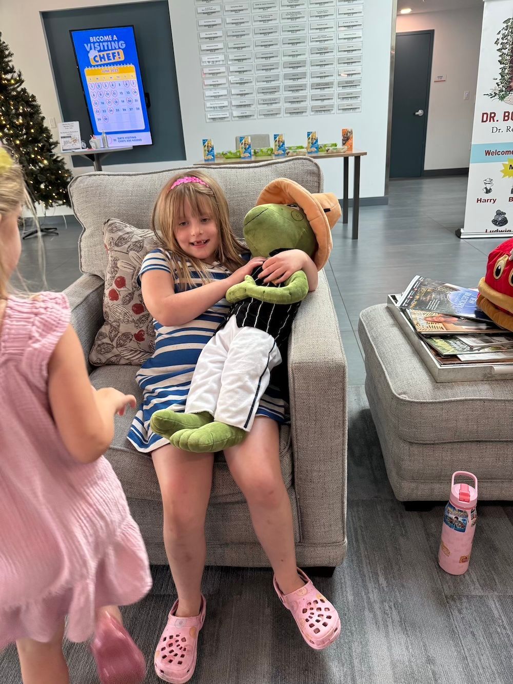 Girl in blue and white striped dress holding a green stuffed animal, pink shoes, in a waiting room.