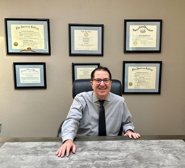 A man is sitting at a desk in front of a wall of framed diplomas.