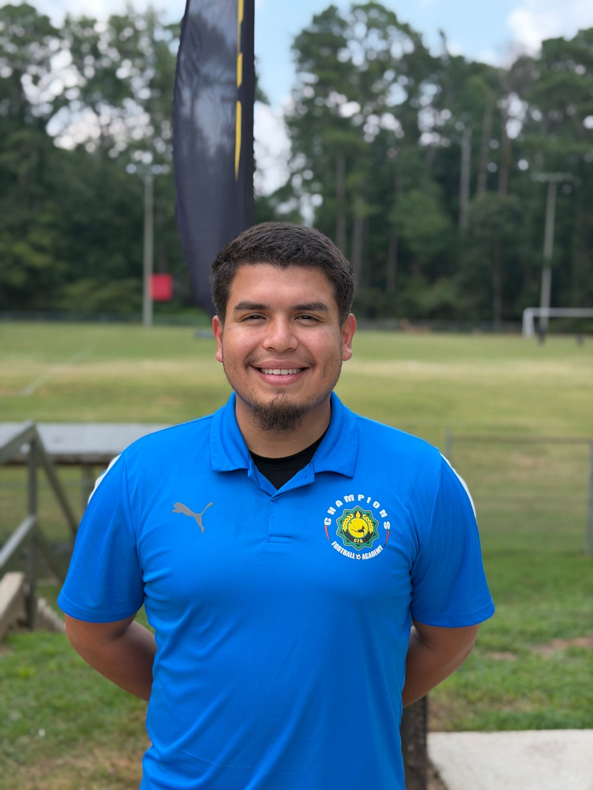 Man in green polo stands on a football field, arms crossed, stadium in background.