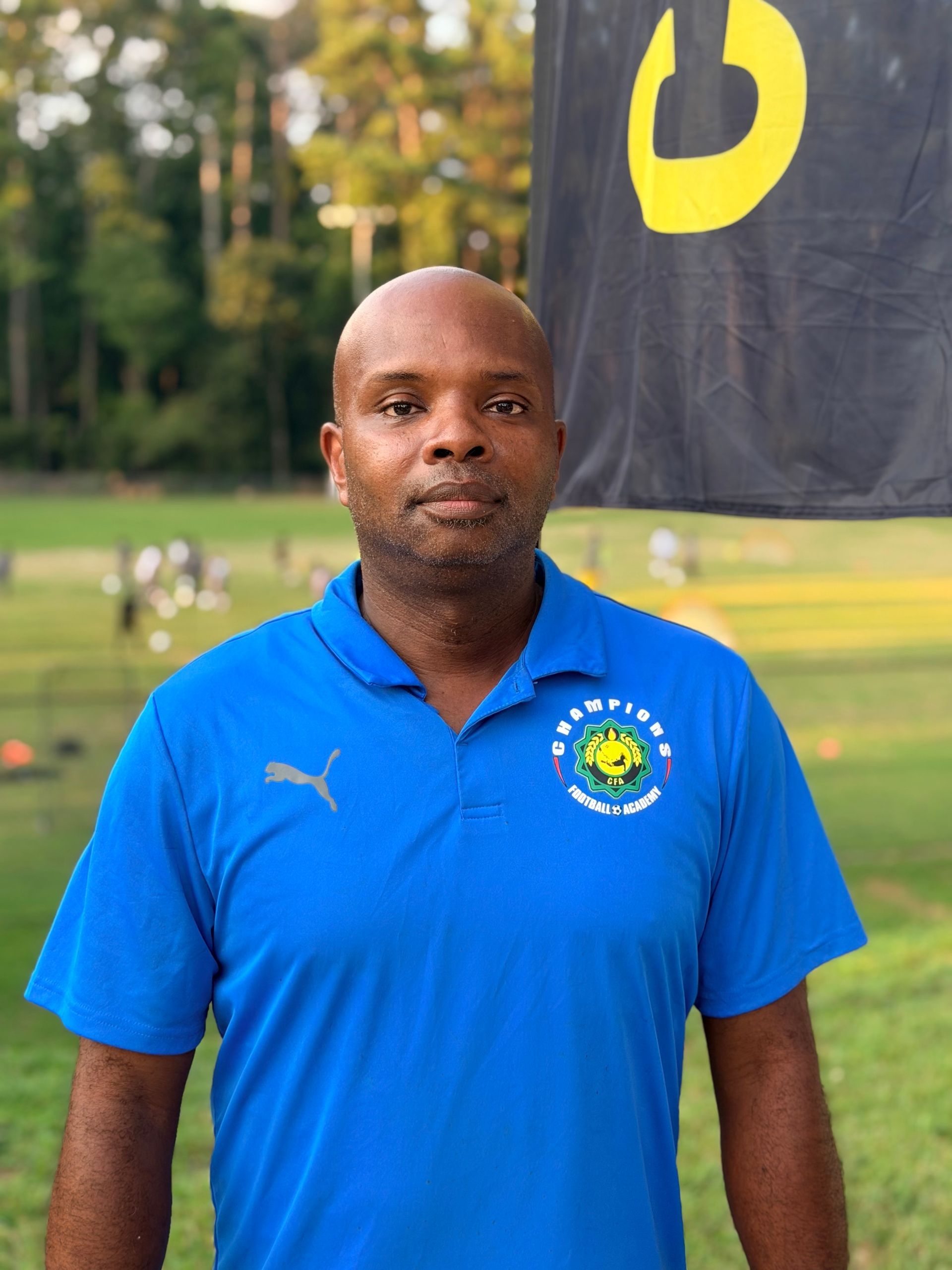 Man in green polo stands on a football field, arms crossed, stadium in background.
