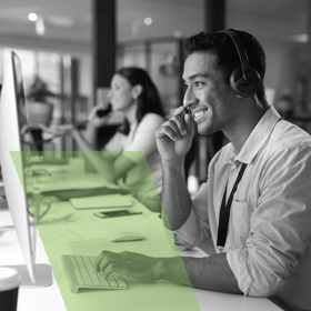 Customer service agent wearing headset working at computer in office, smiling, with a coworker blurred behind him