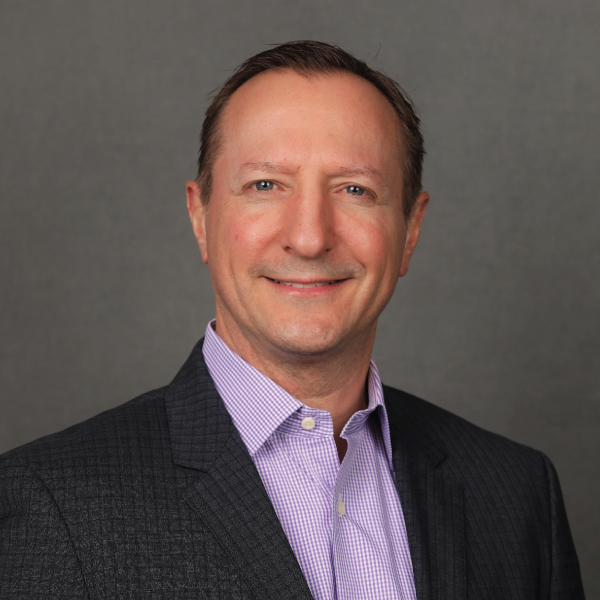 Professional headshot of a man in a dark suit and lavender shirt against a gray background
