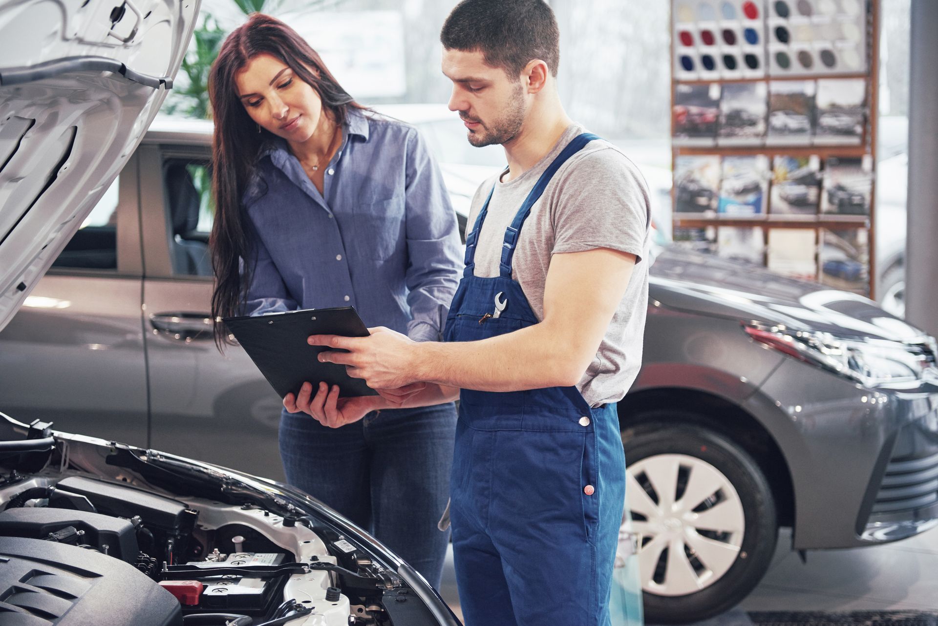 A mechanic in blue overalls and a customer review a checklist in front of an open car hood in a repair shop.
