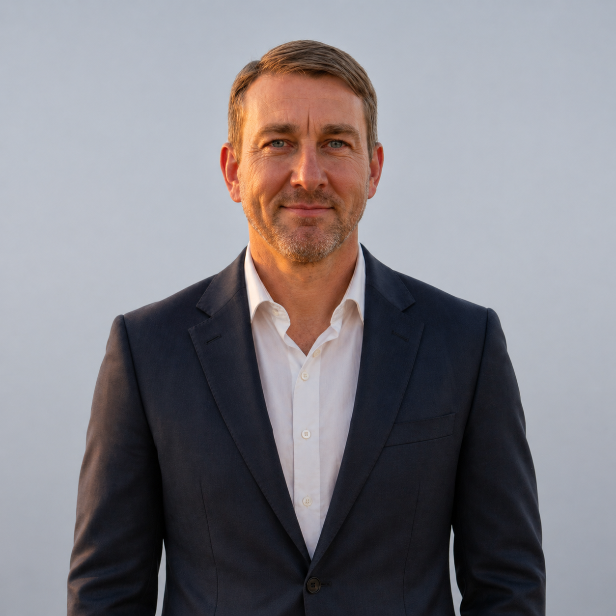 Man in a dark suit and white shirt standing against a plain light background