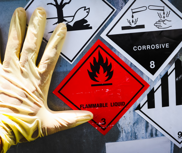 Gloved hand beside hazardous material labels, including red flammable liquid and black corrosive signs.