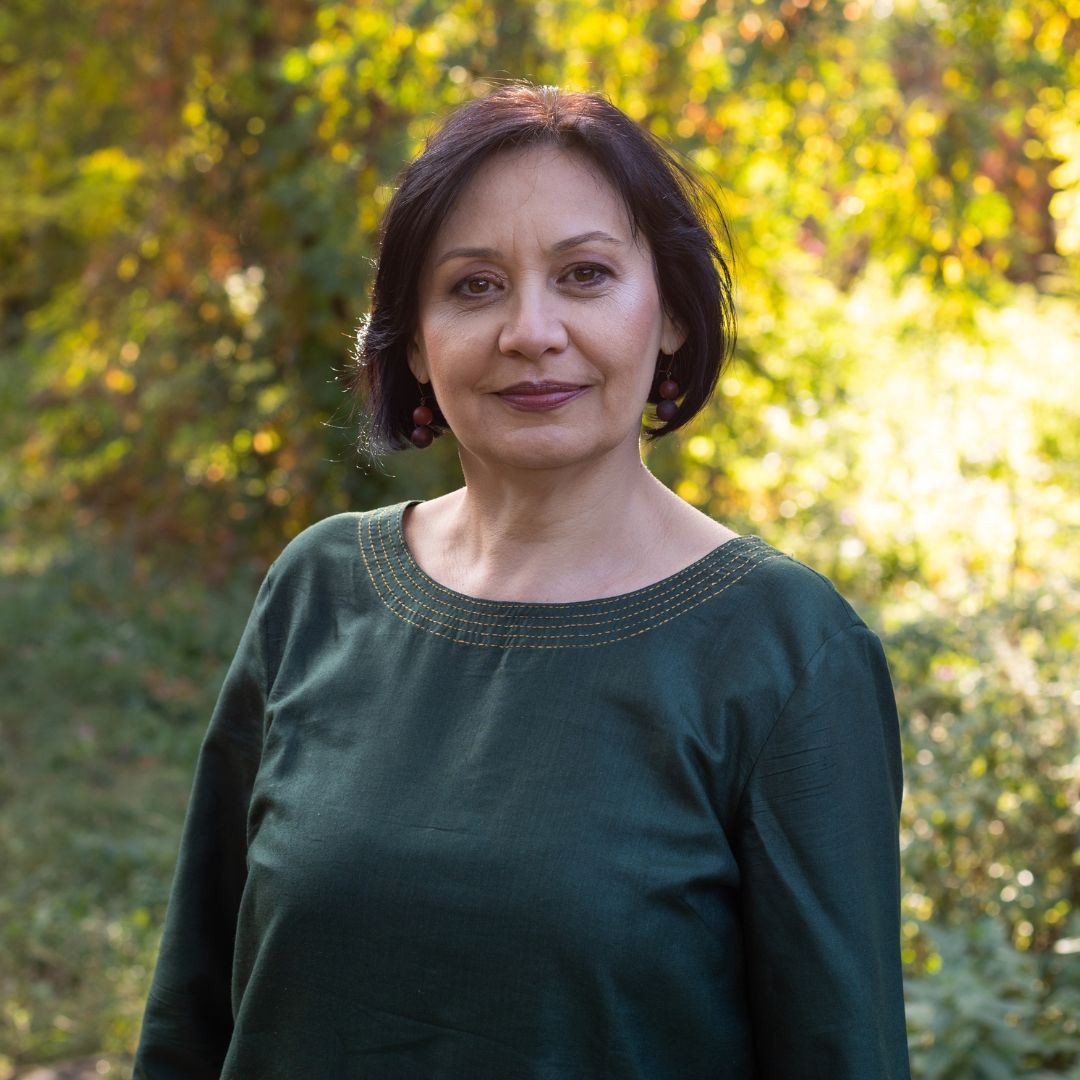 Woman standing outdoors in a green top, with a blurred leafy background and soft sunlight.