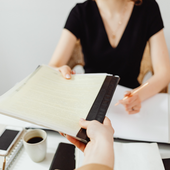 Hands exchanging a binder over a desk with coffee and notebook during a meeting