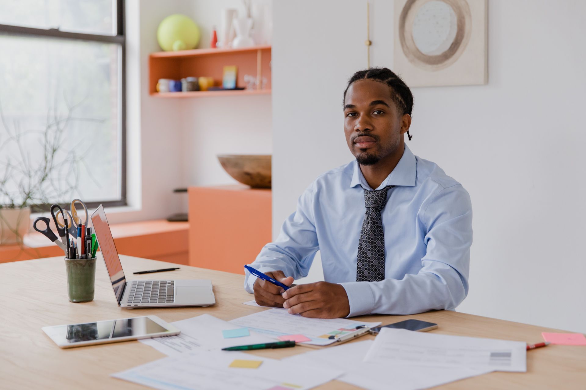 Man seated at a desk in a bright office, looking at papers beside a laptop and coffee cup.
