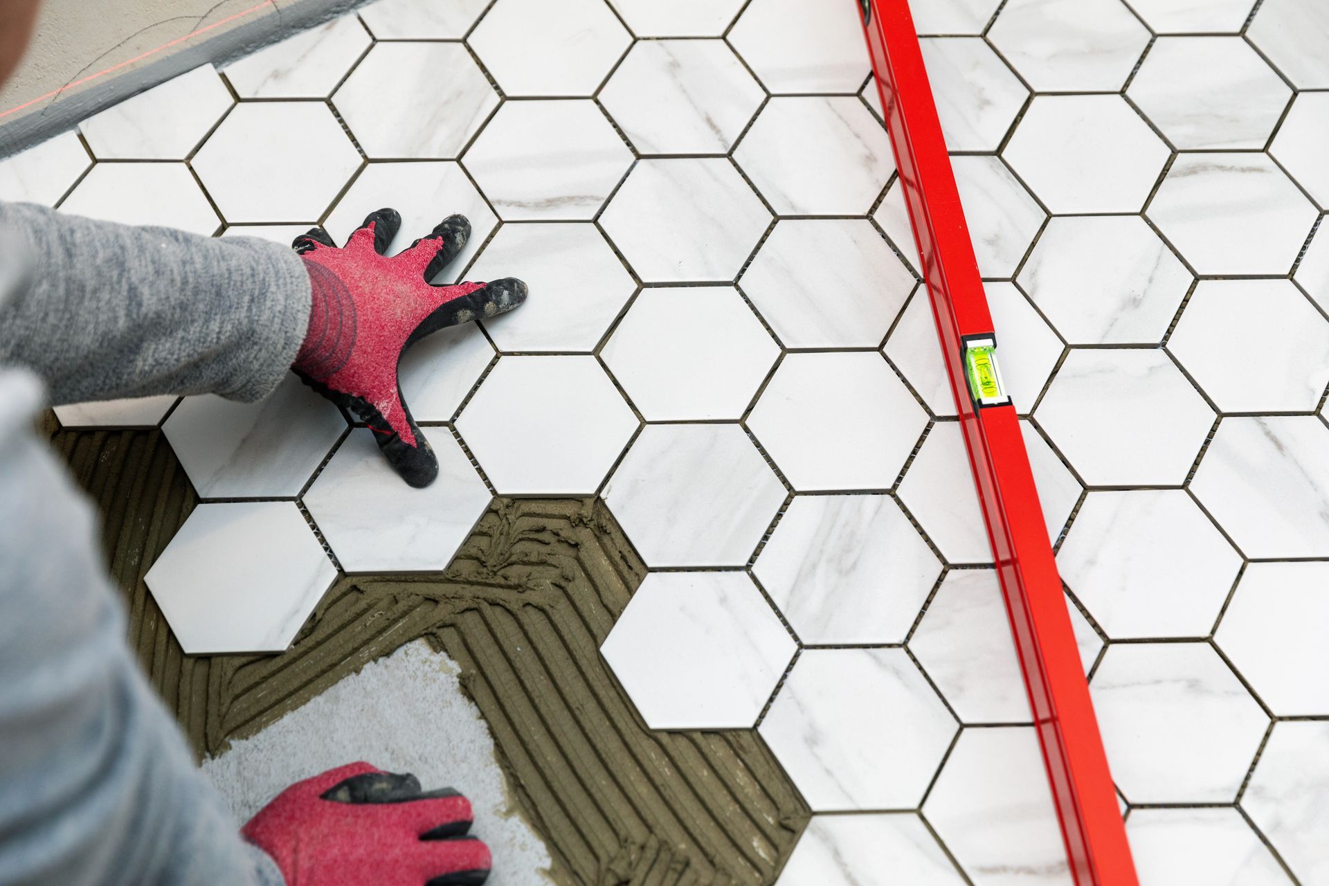 A person wearing gloves lays white hexagonal tiles onto adhesive on a floor, using a red level to ensure alignment.