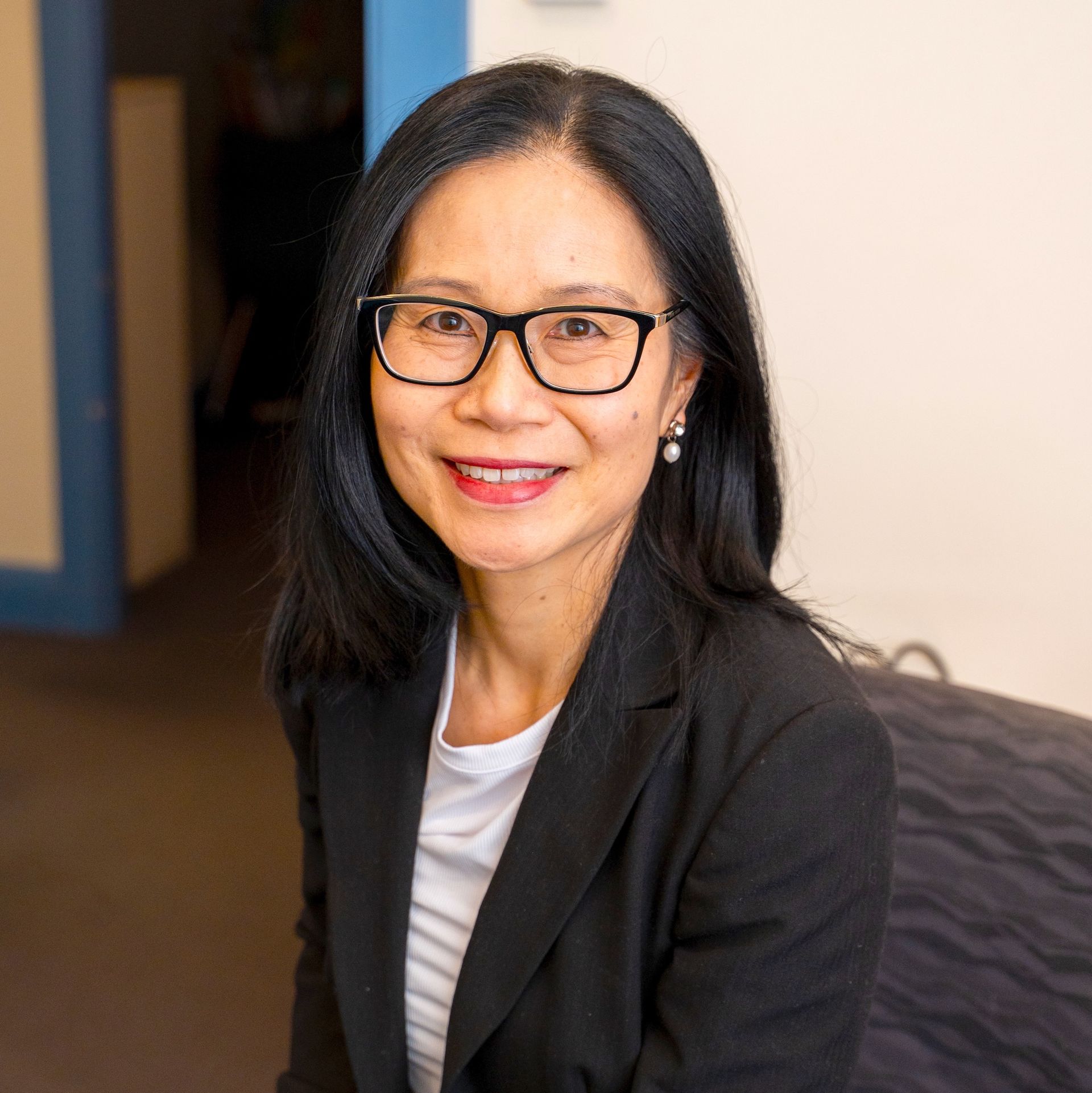 Portrait of a smiling person with glasses in a black blazer, seated indoors against a soft, neutral background