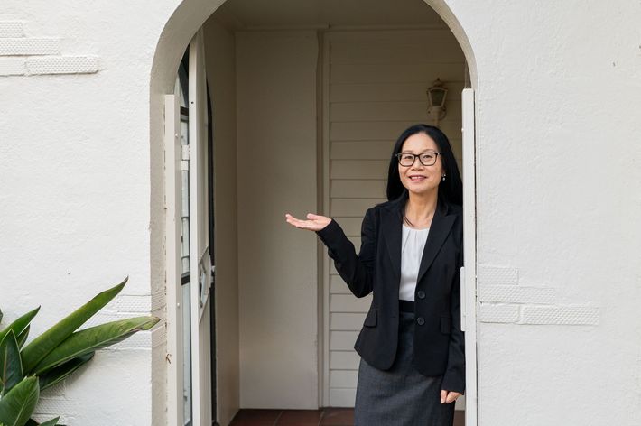 Woman in a black blazer standing in a doorway, gesturing with one hand in a white-walled entryway.