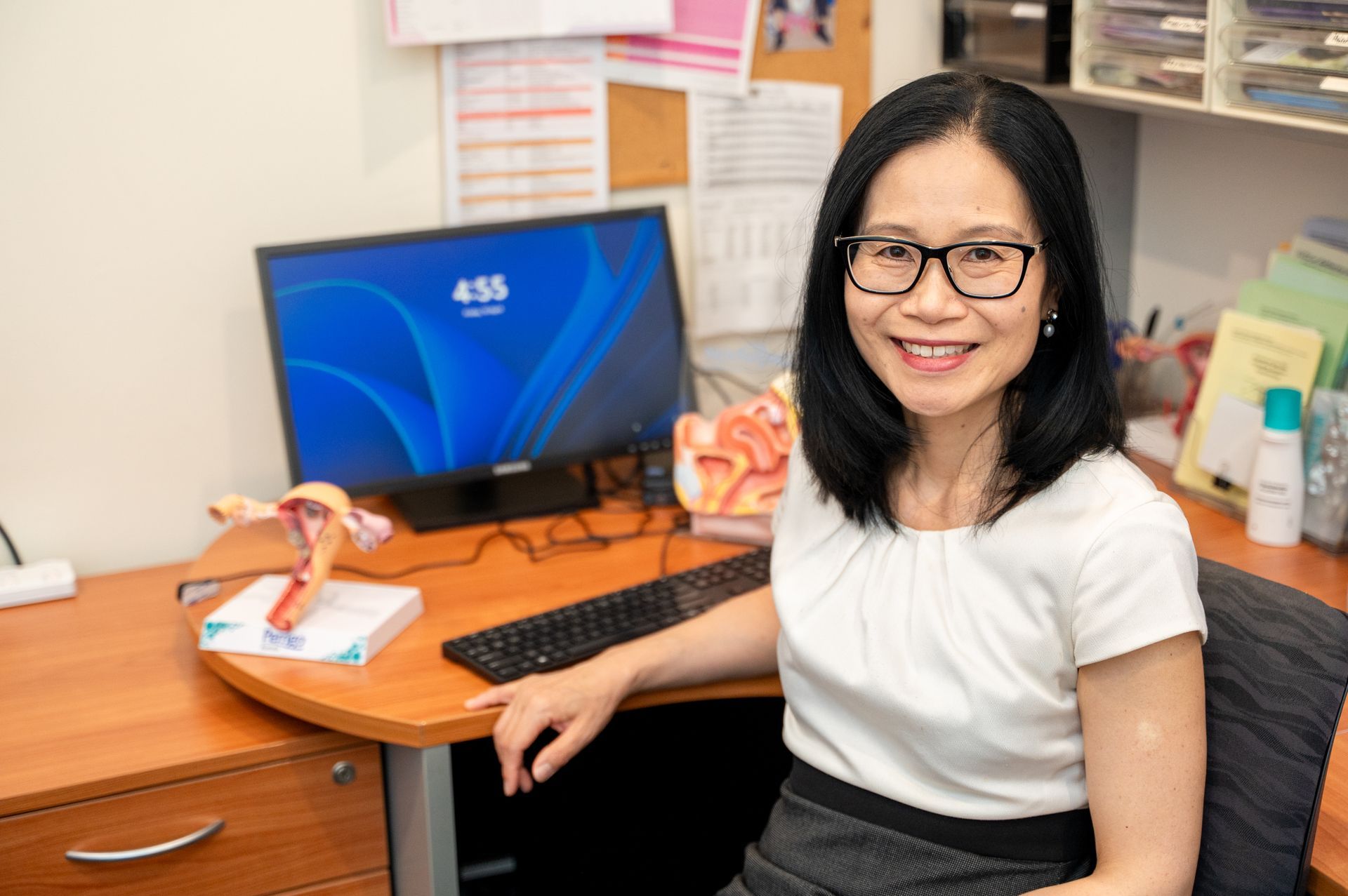 Woman smiling at an office desk with a computer, keyboard, and paperwork.