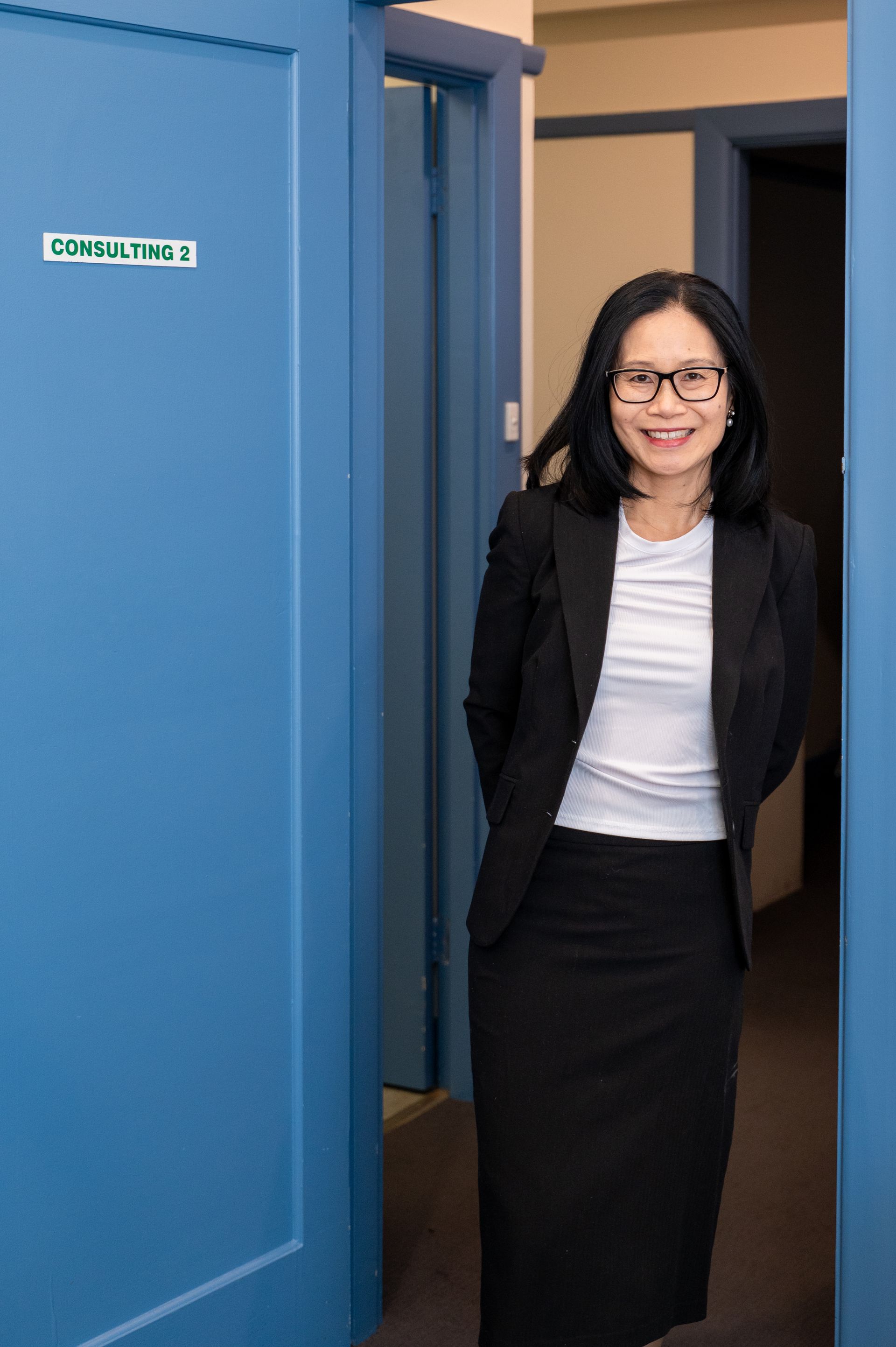 Woman in black blazer stands in a blue doorway, smiling toward the camera.