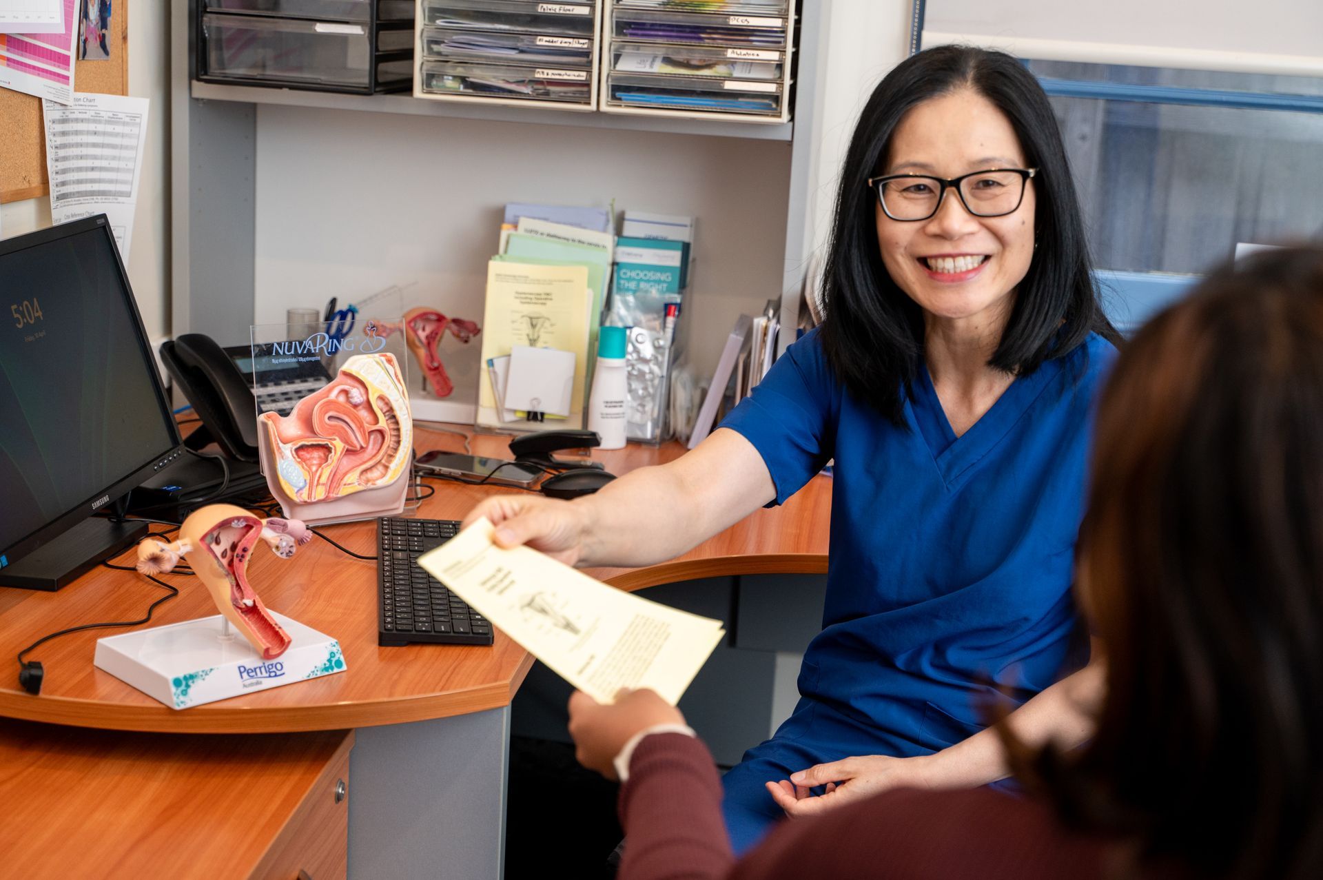 Smiling doctor in blue scrubs handing paperwork to a seated patient in a clinic office