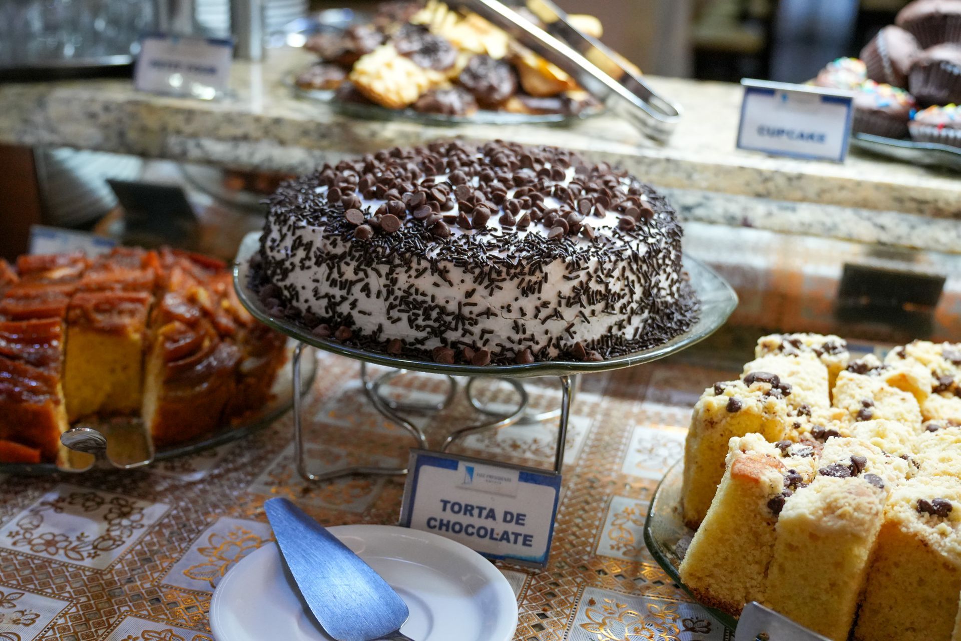 Bolo de chocolate em uma bancada de sobremesas, coberto com raspas de chocolate e ladeado por outros doces.