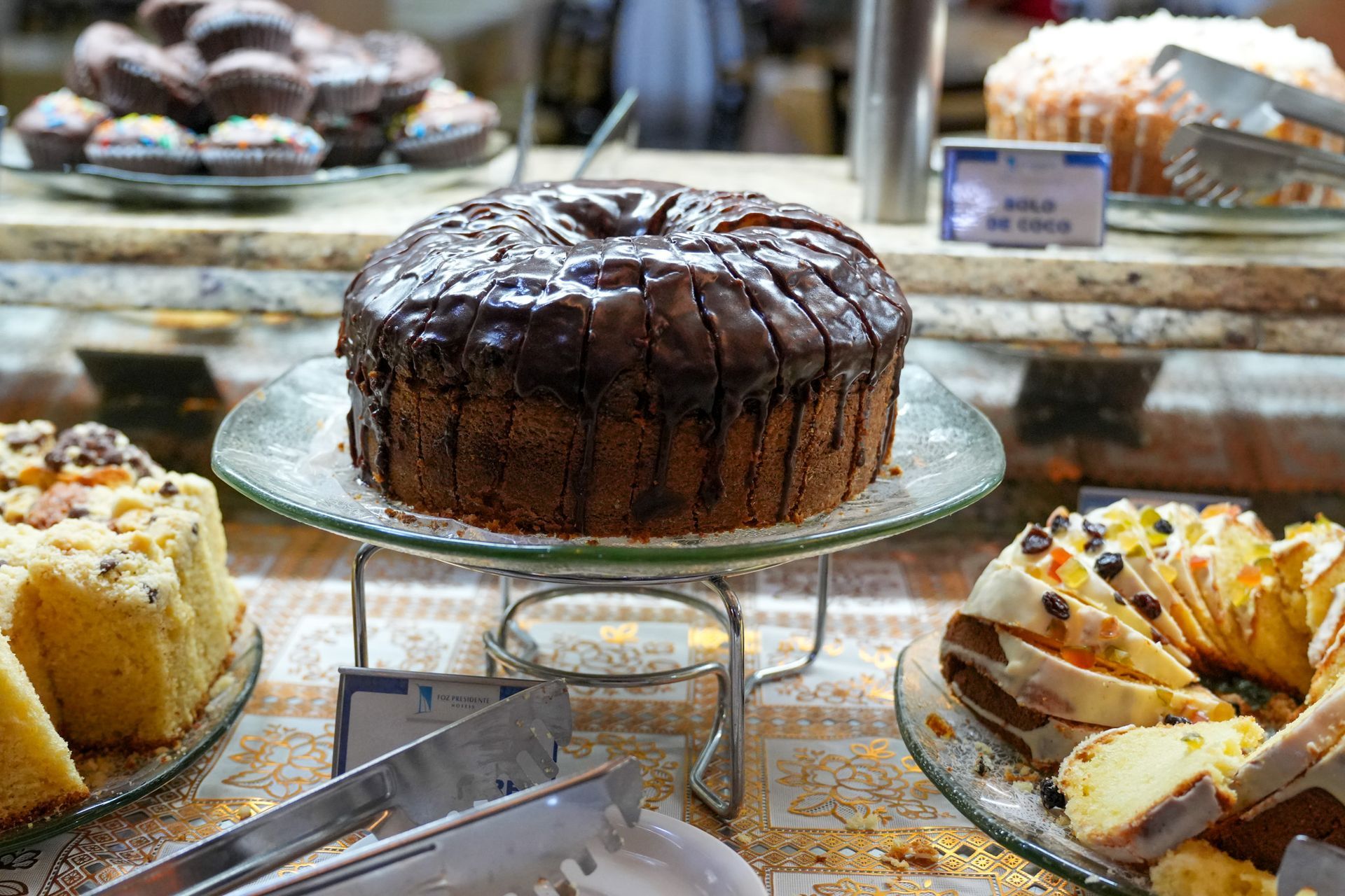 Bolos e doces variados em um balcão de padaria, com um bolo de chocolate no centro de um suporte de vidro.