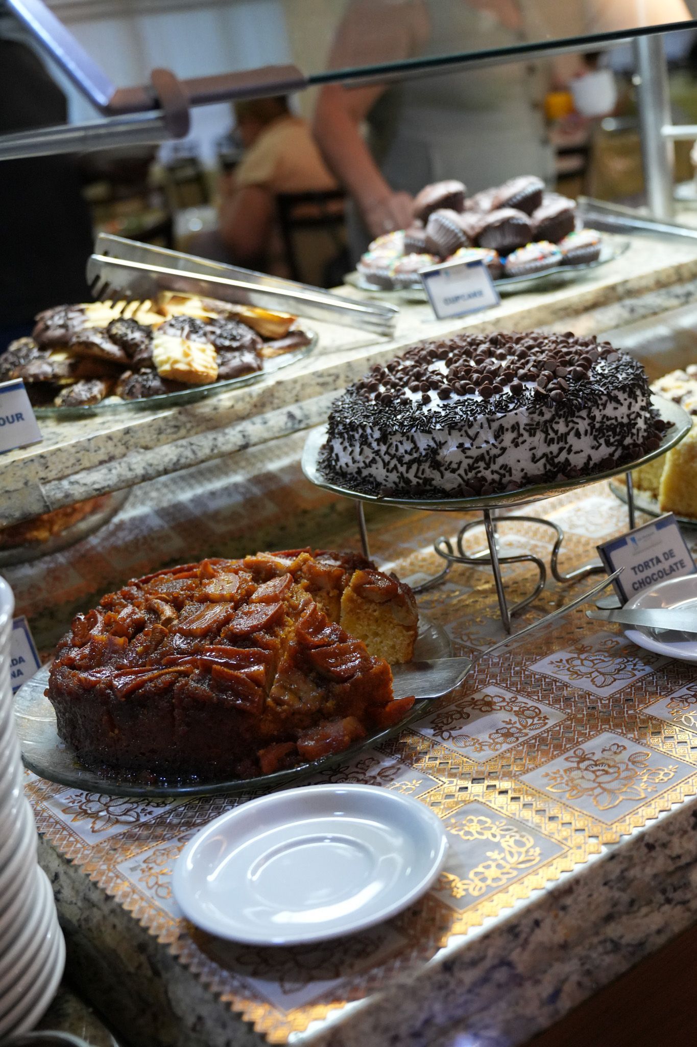 Doces expostos em bandejas atrás do balcão de uma padaria, incluindo um grande bolo com cobertura e um doce com pintinhas.